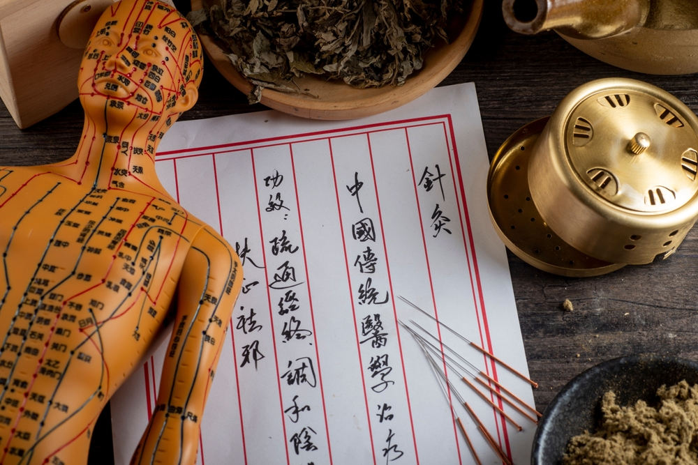 A flat lay of Traditional Chinese Medicine tools and materials on a dark wooden surface, including a yellow acupuncture meridian model, thin acupuncture needles, a brass moxibustion vessel, a bowl of dried herbs, and a handwritten document in Chinese characters.
